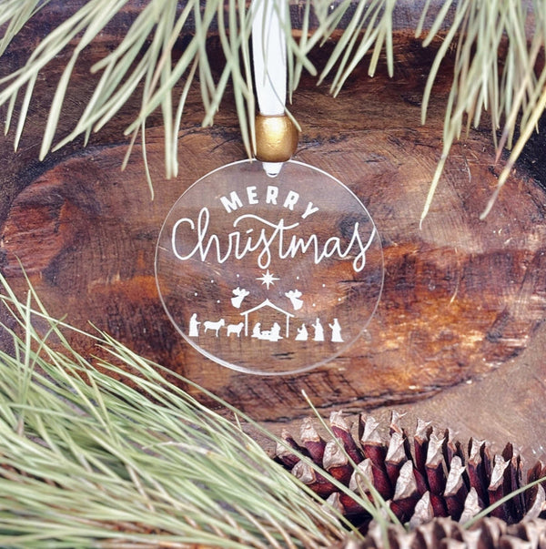 Clear Christmas ornament with 'Merry Christmas' and nativity scene design, hanging on a wooden surface with pine branches.