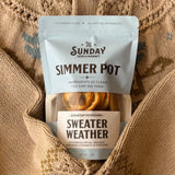 A 2 oz pouch of 'The Sunday Seed of the Market Simmer Pot' in the flavor 'Sweater Weather', nestled inside a beige knit sweater.